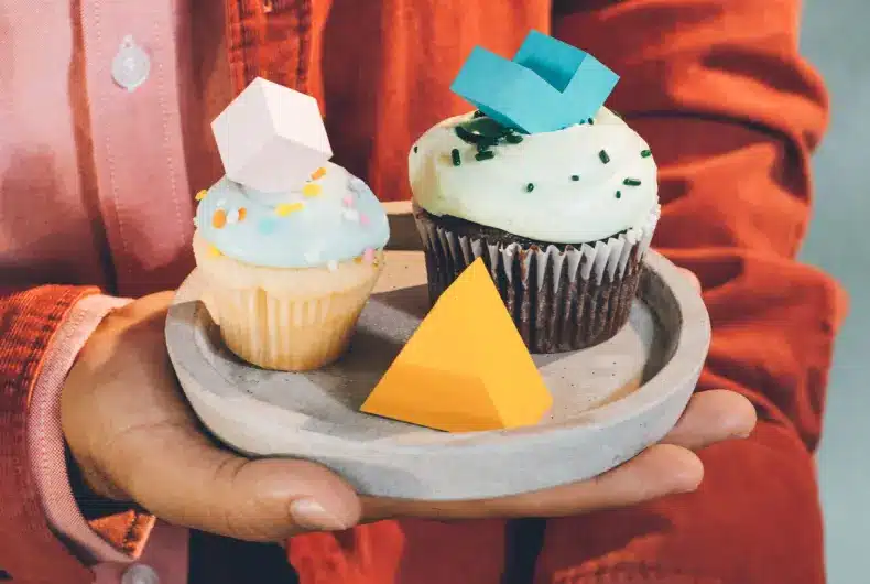 "A man wearing a red jacket holds three cupcakes of varying shapes and colors: black, yellow, and black."